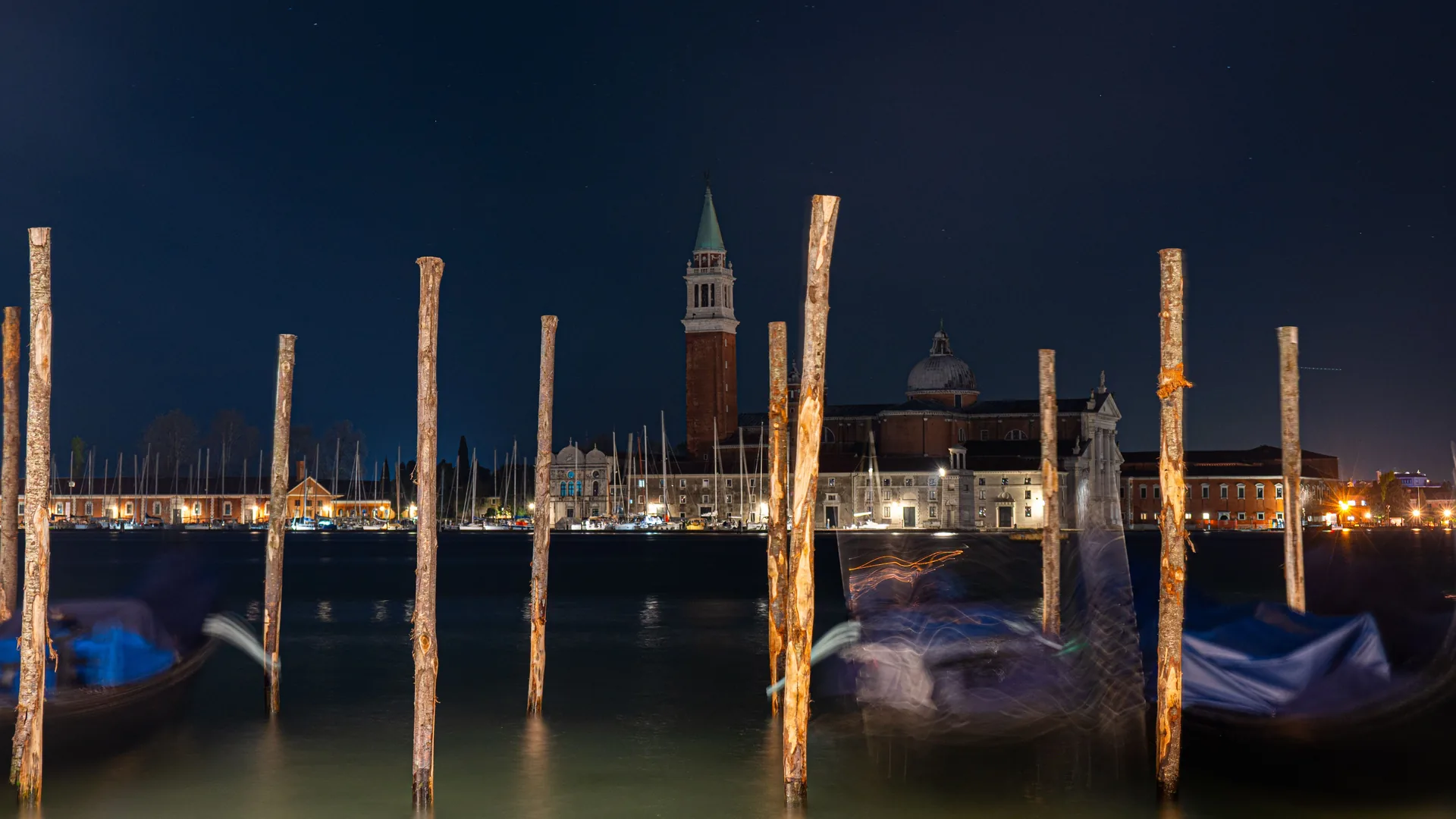 A tall wooden sailing ship on dark water at dusk, lagoon lights on the horizon
